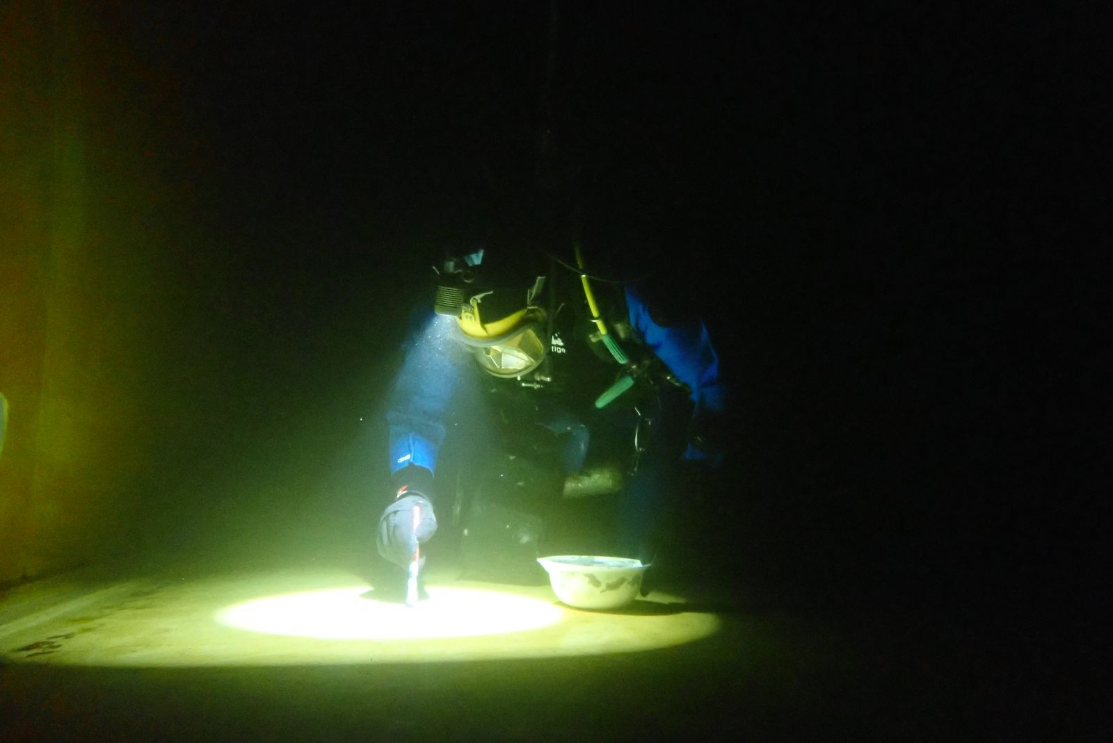 Diver carrying out coating repairs on a reservoir floor under torchlight, with repair material at hand