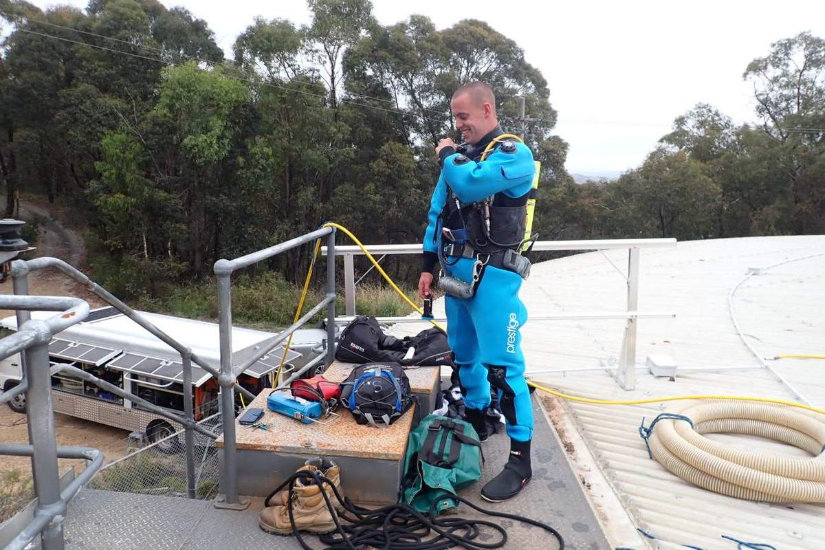 Diver in a blue drysuit preparing equipment on a tank roof platform